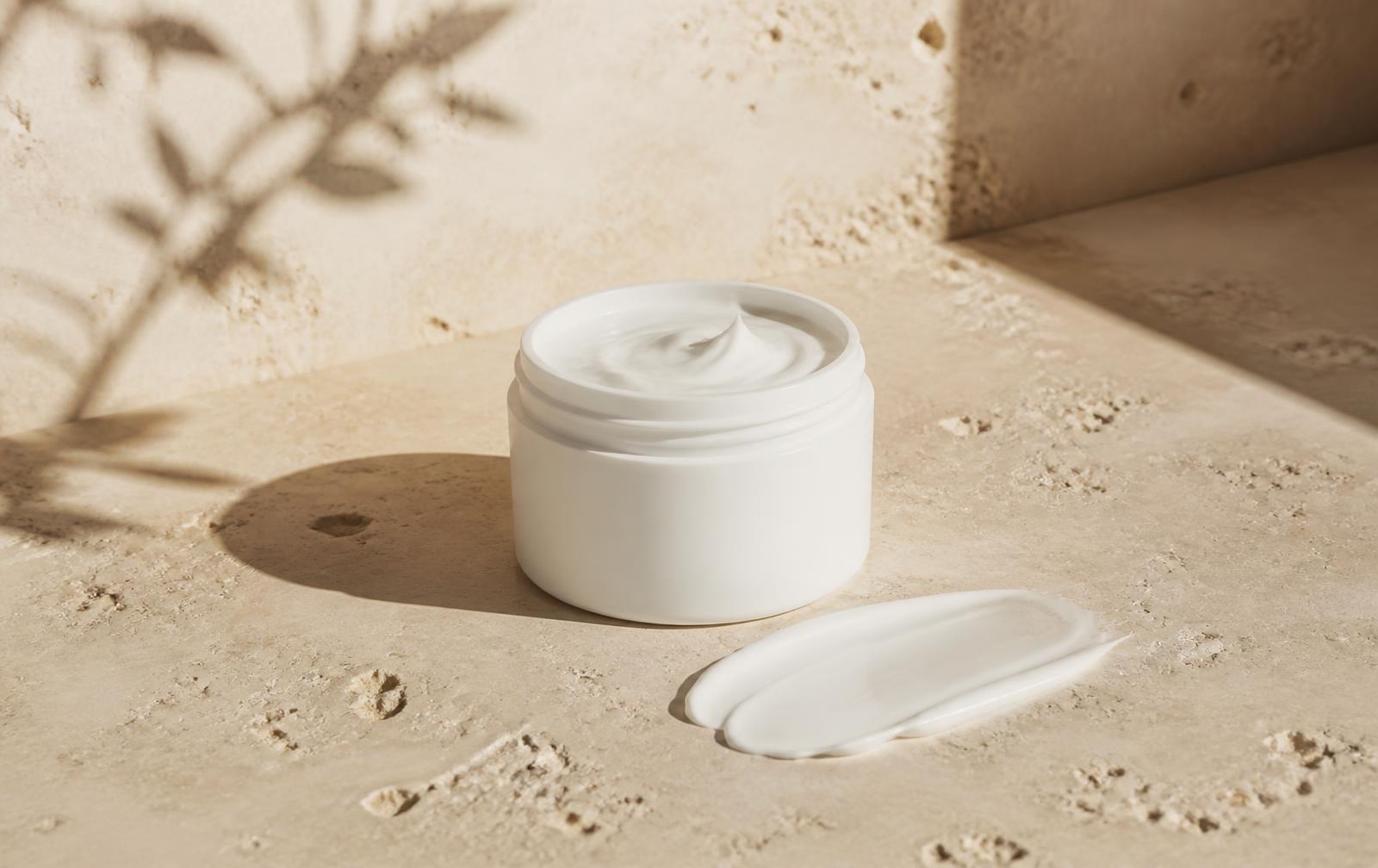 Unbranded white cream jar with a smear of rich cream on warm travertine stone in morning light