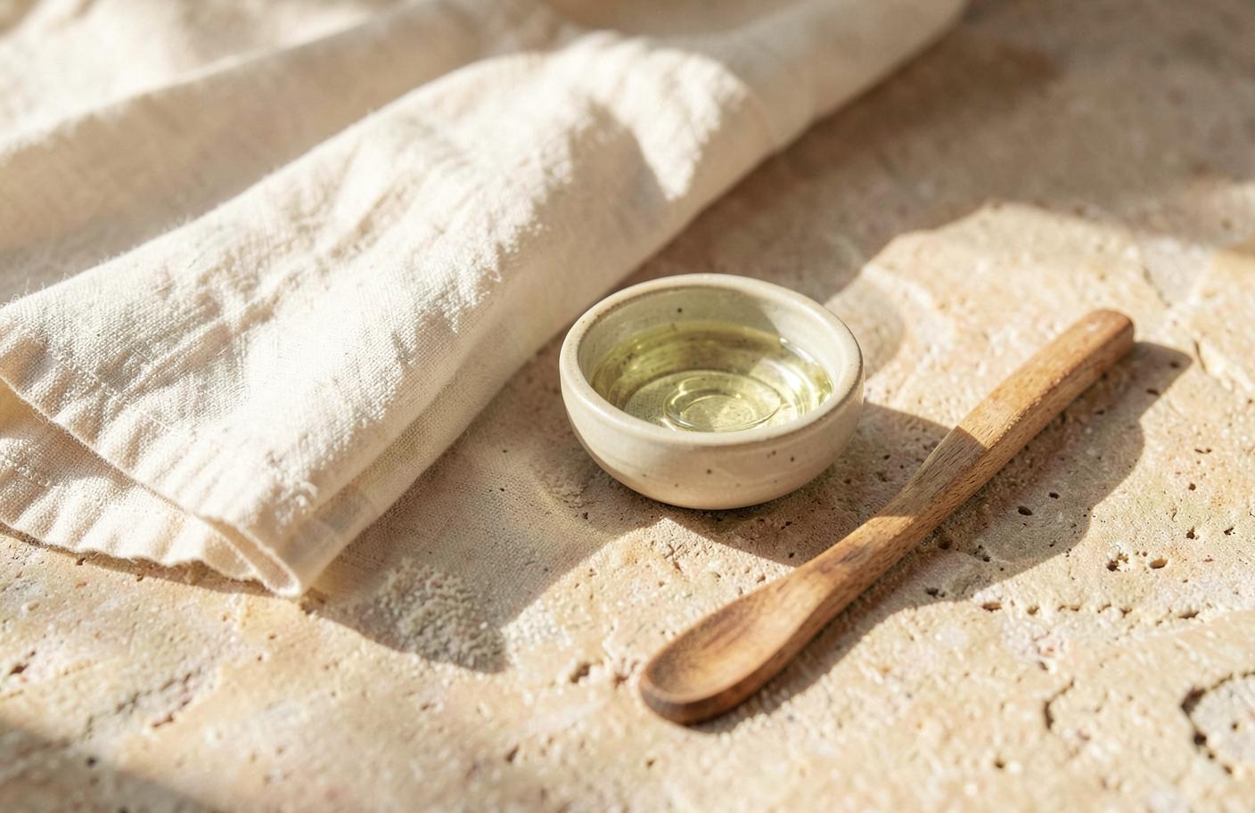 Folded ivory linen cloth beside a small ceramic bowl of clear facial oil on a warm sandstone surface in morning light