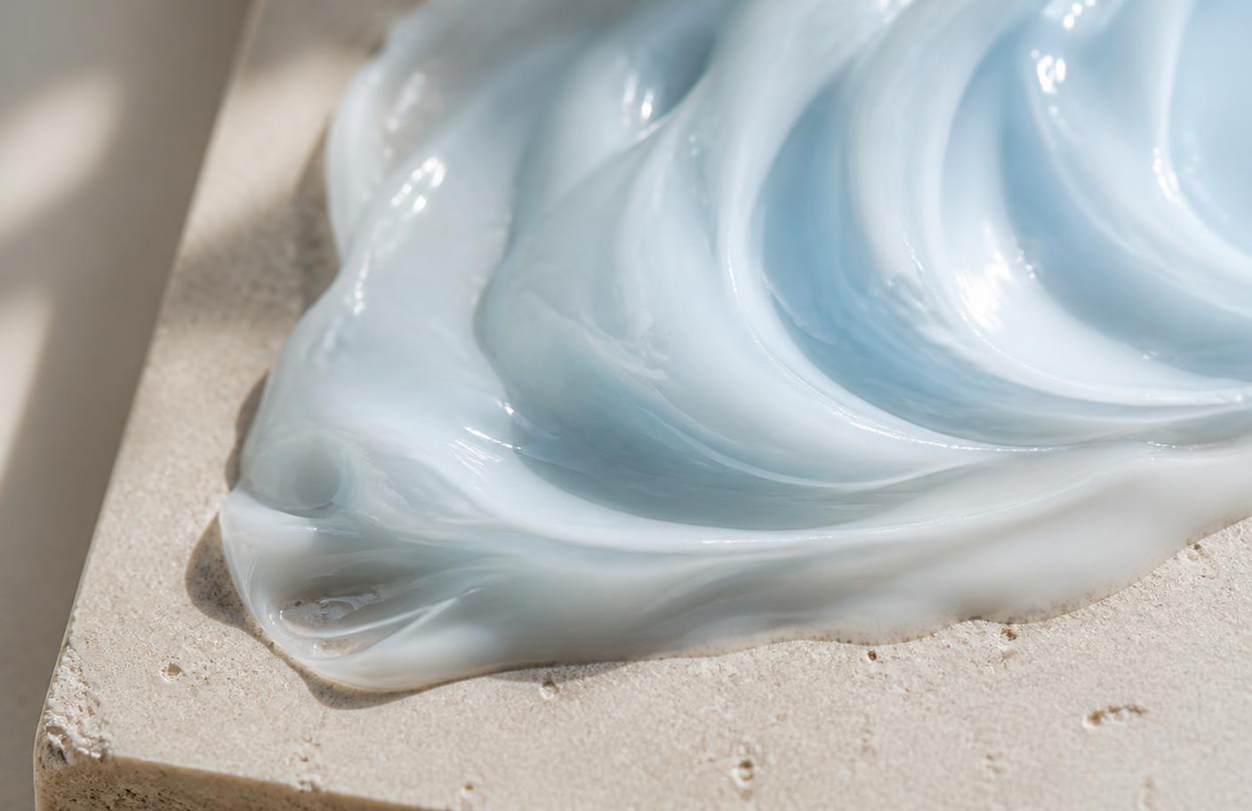 Macro photograph of a pale blue-tinted gel cream swirled on a warm stone tile, showing peptide moisturiser texture