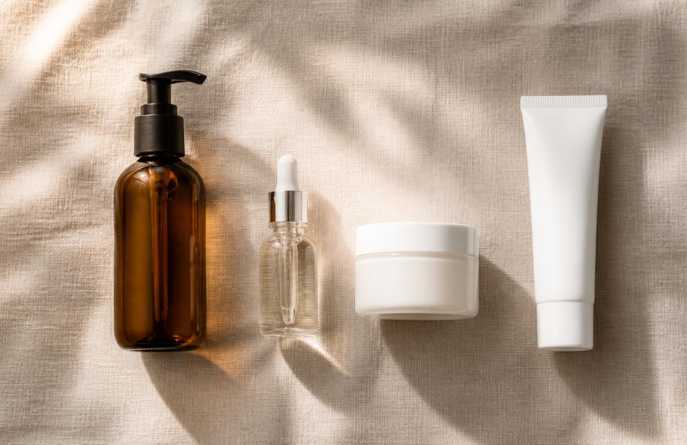 Overhead flat lay of a simple four-step skincare routine on linen: cleanser, serum, moisturiser and sunscreen
